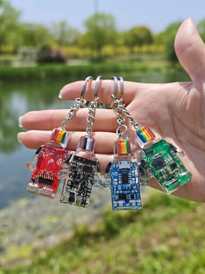 May include: Four colorful robot-shaped keychains. Each keychain features a clear body with internal circuit board details and a rainbow-colored top. The keychains are red, black, blue, and green, attached to silver keyrings.