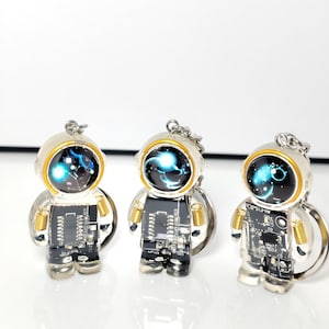 May include: Three astronaut-shaped keychains featuring black circuit board bodies with gold-colored coil details on the arms. Each figure has a round helmet with a dark, glossy visor displaying a glowing blue nebula-like graphic and a silver ring.