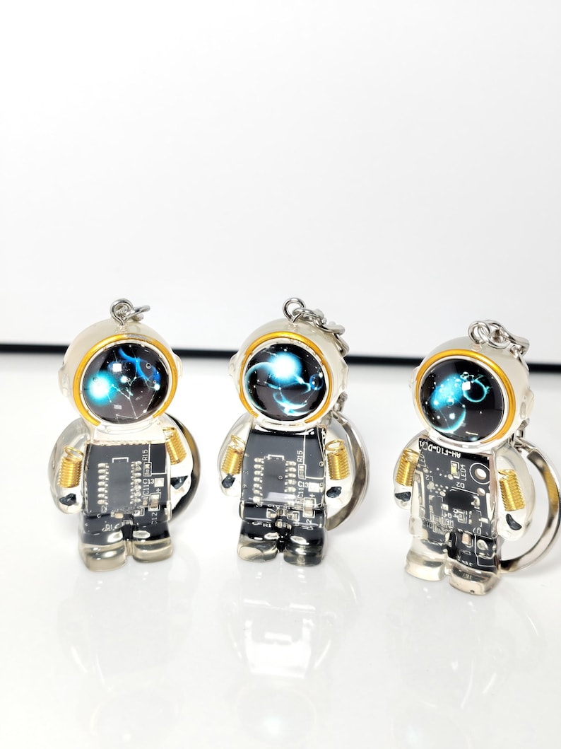 May include: Three astronaut-shaped keychains featuring black circuit board bodies with gold-colored coil details on the arms. Each figure has a round helmet with a dark, glossy visor displaying a glowing blue nebula-like graphic and a silver ring.