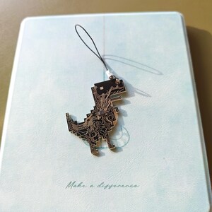 May include: A black and gold dinosaur charm with a circuit board design, attached to a black string. The charm is resting on a light blue notebook with the words "Make a difference" printed on it.