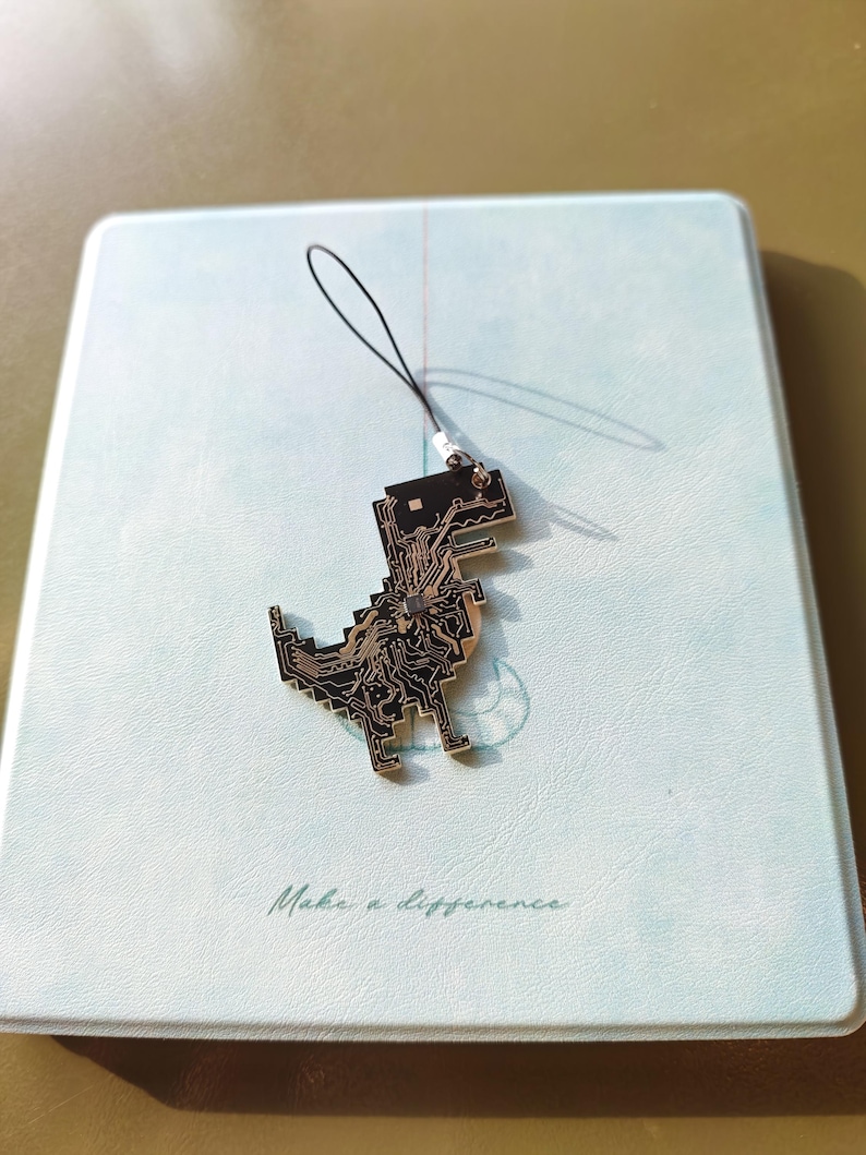 May include: A black and gold dinosaur charm with a circuit board design, attached to a black string. The charm is resting on a light blue notebook with the words "Make a difference" printed on it.