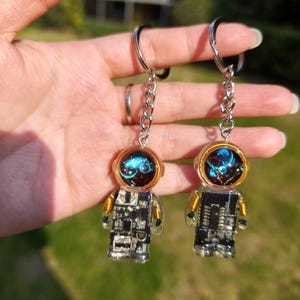 May include: Two astronaut-shaped keychains featuring clear resin bodies embedded with black and silver electronic circuit board components, gold wire coils at the shoulders, and helmets with glowing blue nebula-like patterns inside.