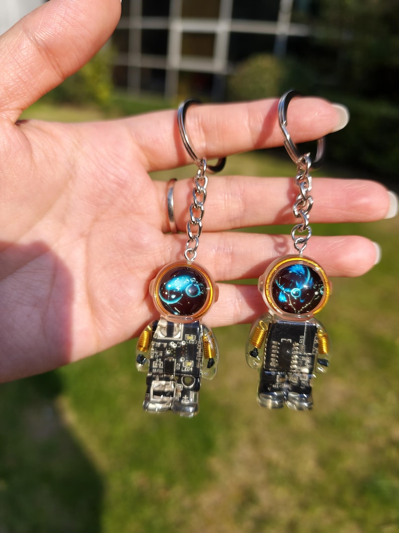 May include: Two astronaut-shaped keychains featuring clear resin bodies embedded with black and silver electronic circuit board components, gold wire coils at the shoulders, and helmets with glowing blue nebula-like patterns inside.