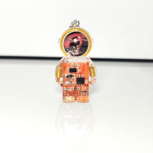 May include: A clear resin astronaut pendant featuring an orange circuit board interior with visible electronic components, gold-colored wire coils at the shoulders, and a helmet visor displaying a dark space-themed graphic with stars.
