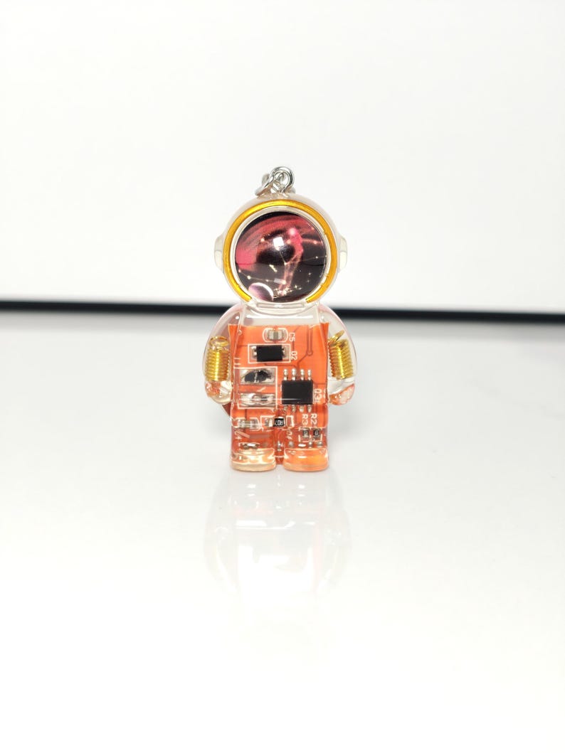 May include: A clear resin astronaut pendant featuring an orange circuit board interior with visible electronic components, gold-colored wire coils at the shoulders, and a helmet visor displaying a dark space-themed graphic with stars.
