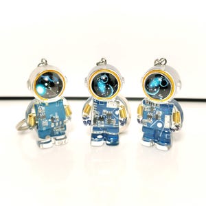 May include: Three astronaut-shaped keychains made of clear resin. Each figure features a blue circuit board torso, gold-colored coil accents at the shoulders, and a helmet with a dark, circular visor displaying a blue and white nebula graphic.