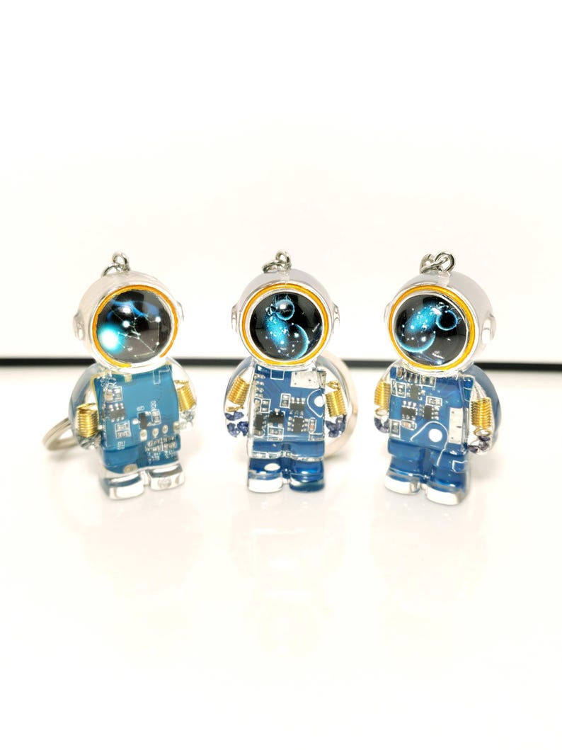 May include: Three astronaut-shaped keychains made of clear resin. Each figure features a blue circuit board torso, gold-colored coil accents at the shoulders, and a helmet with a dark, circular visor displaying a blue and white nebula graphic.
