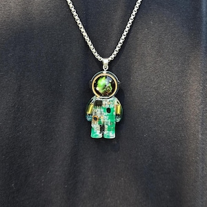 May include: A silver-toned chain necklace featuring a pendant shaped like an astronaut. The pendant is encased in clear resin and contains a green circuit board with visible electronic components and a helmet visor displaying a swirling green nebula.