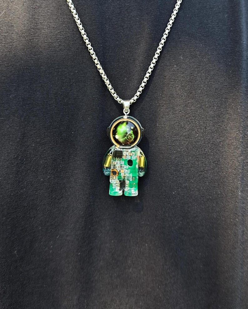 May include: A silver-toned chain necklace featuring a pendant shaped like an astronaut. The pendant is encased in clear resin and contains a green circuit board with visible electronic components and a helmet visor displaying a swirling green nebula.