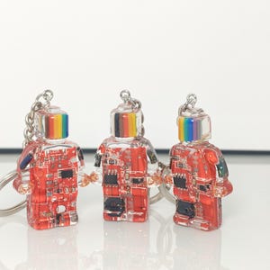 May include: Three clear robot-shaped keychains with red circuit board designs inside. Each robot has a rainbow-colored top and a silver keychain ring. The keychains are on a white surface.