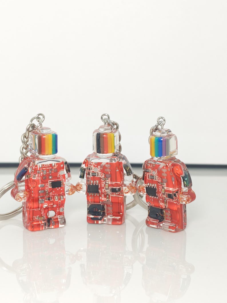May include: Three clear robot-shaped keychains with red circuit board designs inside. Each robot has a rainbow-colored top and a silver keychain ring. The keychains are on a white surface.