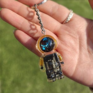 May include: A keychain featuring a clear resin astronaut figure with a black circuit board torso and gold-colored wire coils at the shoulders. The helmet visor is dark with blue and white light reflections, and the figure hangs from a silver metal ring.