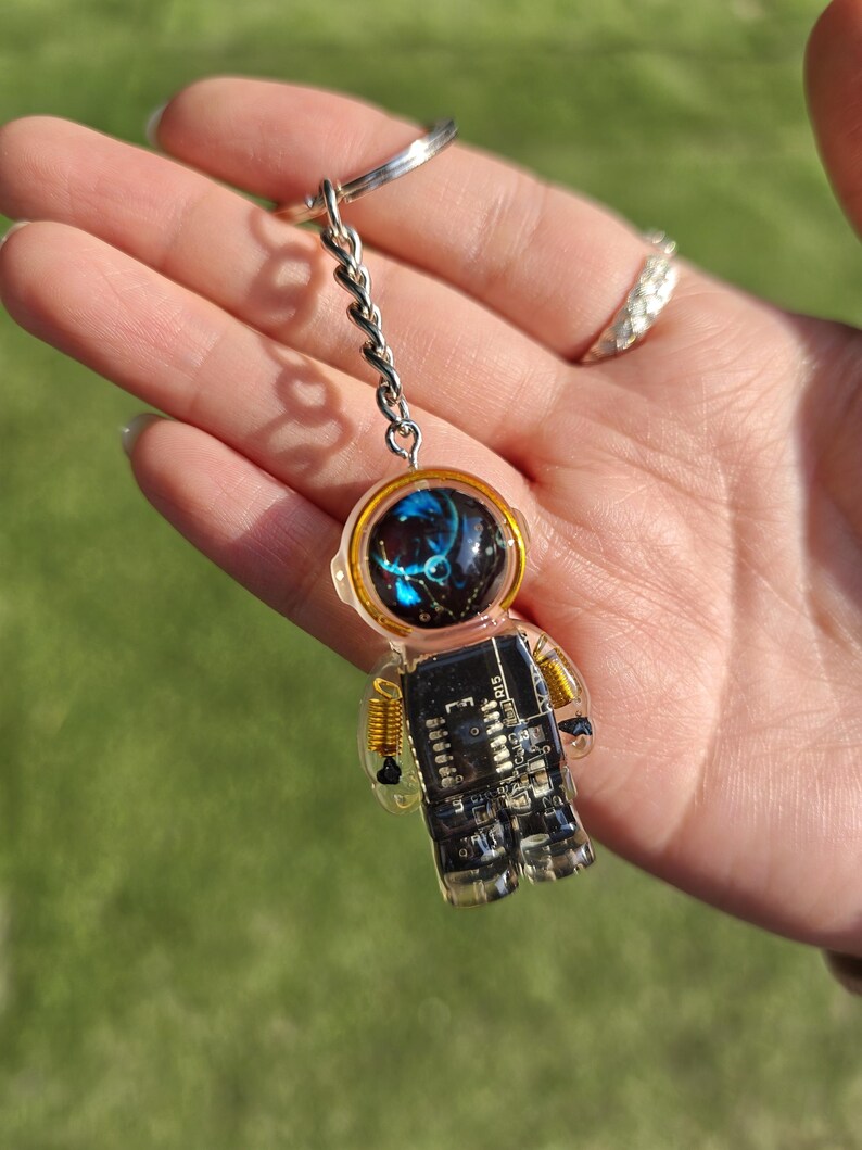 May include: A keychain featuring a clear resin astronaut figure with a black circuit board torso and gold-colored wire coils at the shoulders. The helmet visor is dark with blue and white light reflections, and the figure hangs from a silver metal ring.