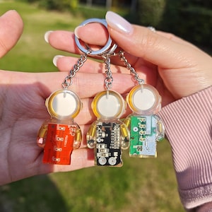 May include: Three astronaut-shaped keychains made of clear resin, each featuring a circular white helmet and a rectangular circuit board body in red, black, or green. The figures are attached to silver metal chains and a split ring for keys.