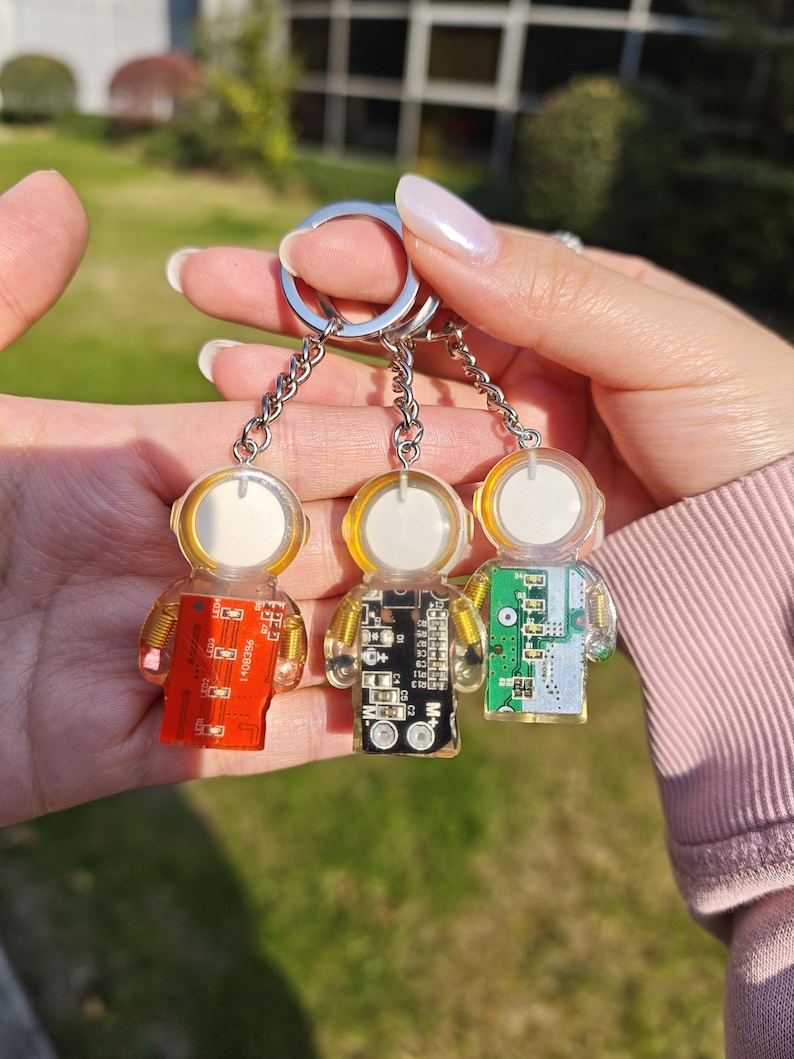 May include: Three astronaut-shaped keychains made of clear resin, each featuring a circular white helmet and a rectangular circuit board body in red, black, or green. The figures are attached to silver metal chains and a split ring for keys.