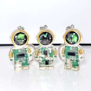 May include: Three astronaut-shaped keychains made of clear resin, each containing a green circuit board with visible electronic components and gold-colored springs at the shoulders. The helmets feature a black dome with a glowing green graphic.