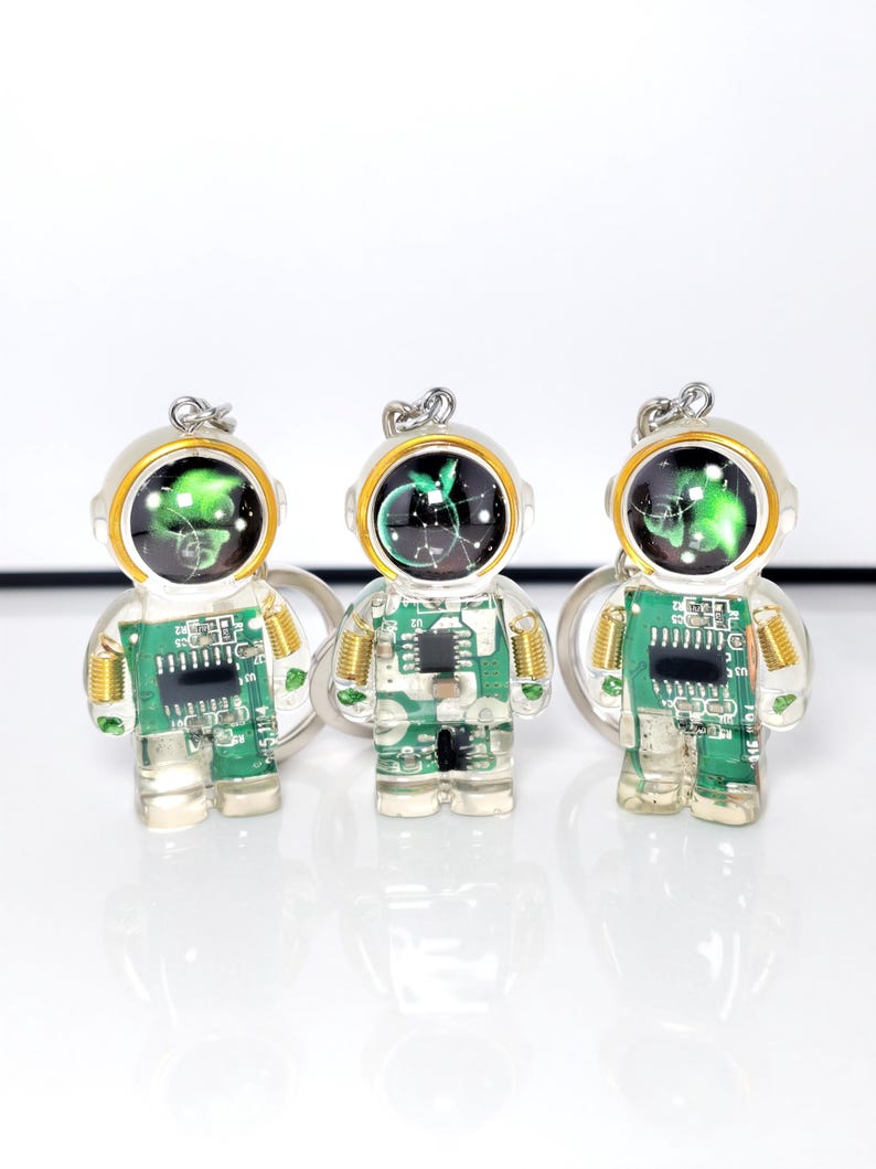 May include: Three astronaut-shaped keychains made of clear resin, each containing a green circuit board with visible electronic components and gold-colored springs at the shoulders. The helmets feature a black dome with a glowing green graphic.