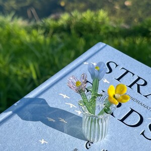 May include: A small glass vase with three colorful glass flowers, resting on a light blue book. The flowers are pink, yellow, and blue. The book has the word "STR" visible on the cover.