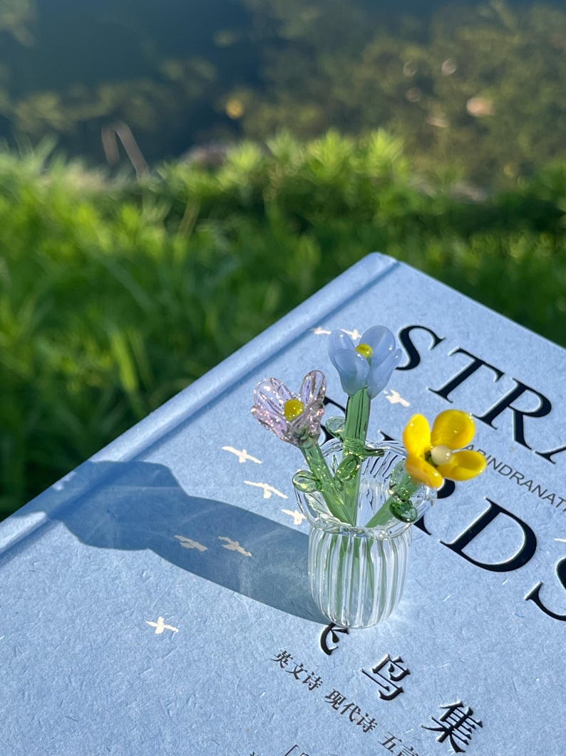 May include: A small glass vase with three colorful glass flowers, resting on a light blue book. The flowers are pink, yellow, and blue. The book has the word "STR" visible on the cover.