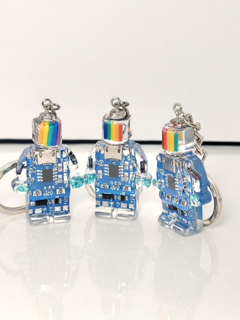 May include: Three clear robot keychains with rainbow-colored heads and blue circuit board bodies. Each keychain has a silver ring and small blue accents. The robots stand on a white surface.