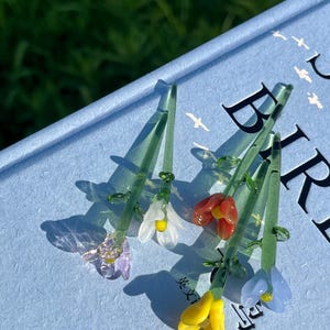 May include: A collection of delicate glass flower sculptures in various colors, including purple, white, red, yellow, and blue. Each flower has a green stem and is resting on a light blue surface. The flowers are intricately detailed.