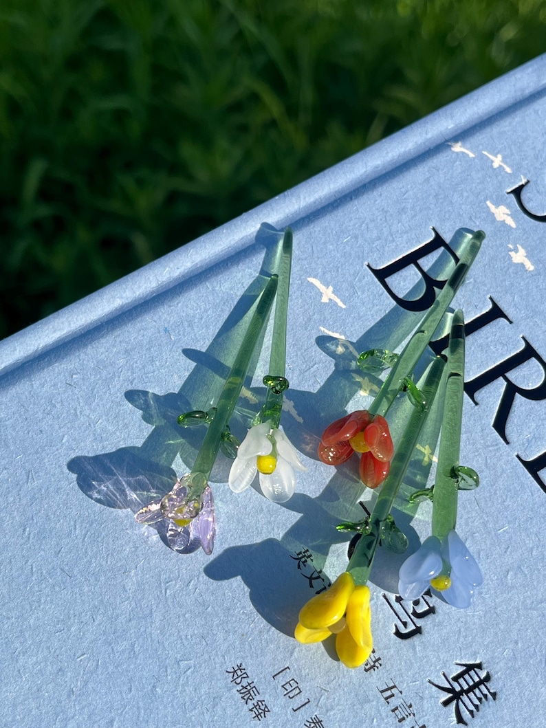 May include: A collection of delicate glass flower sculptures in various colors, including purple, white, red, yellow, and blue. Each flower has a green stem and is resting on a light blue surface. The flowers are intricately detailed.