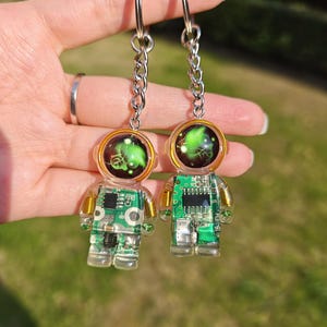 May include: Two astronaut-shaped keychains made of clear resin, each featuring an embedded green circuit board with visible electronic components and gold-colored wire coils. The helmets contain a swirling green and black nebula-like design.