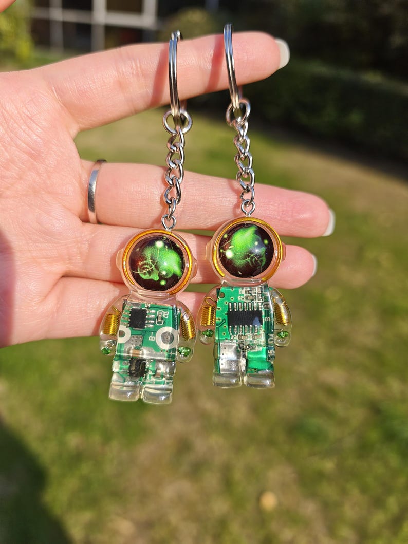 May include: Two astronaut-shaped keychains made of clear resin, each featuring an embedded green circuit board with visible electronic components and gold-colored wire coils. The helmets contain a swirling green and black nebula-like design.