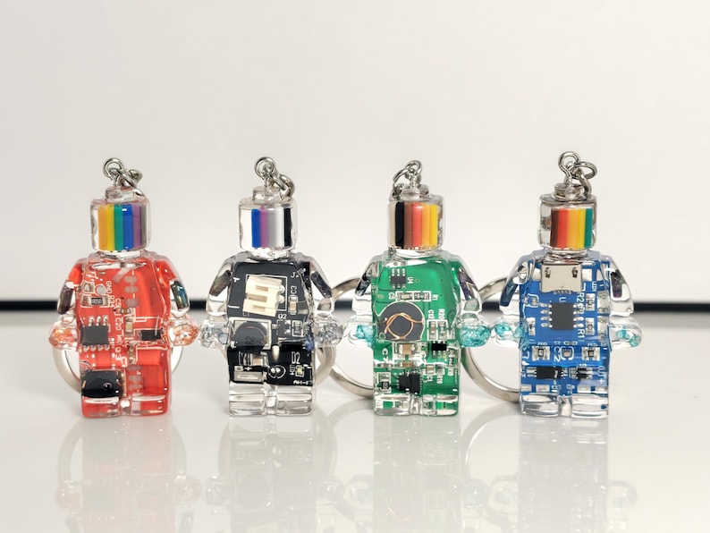 May include: Four robot-shaped keychains with clear bodies and circuit board designs. Each robot has a different colored body: red, black, green, and blue. The robots have rainbow-colored or silver tops and are attached to silver keyrings.
