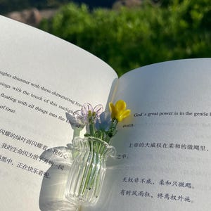May include: A small, clear glass vase with a ruffled rim, holding delicate glass flowers in yellow, purple, and blue. The vase sits on an open book with text in English and Chinese, with a blurred green background.