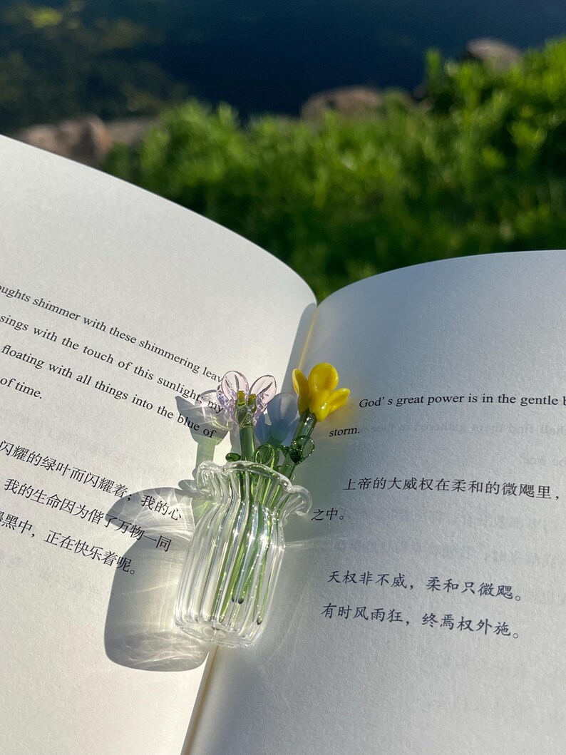 May include: A small, clear glass vase with a ruffled rim, holding delicate glass flowers in yellow, purple, and blue. The vase sits on an open book with text in English and Chinese, with a blurred green background.