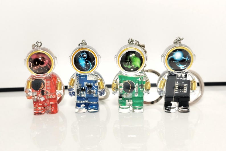 May include: Four astronaut-shaped keychains made of clear resin, each featuring internal electronic circuit board patterns in red, blue, green, and black. Each figure has a metallic ring, gold-toned coil details on the shoulders, and a helmet visor.