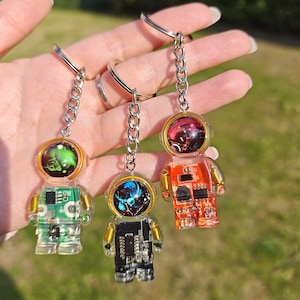 May include: Three astronaut-shaped keychains made of clear resin, each featuring a unique internal circuit board design in green, black, and orange. Each figure has a metallic helmet visor and is attached to a silver-toned metal chain and ring.