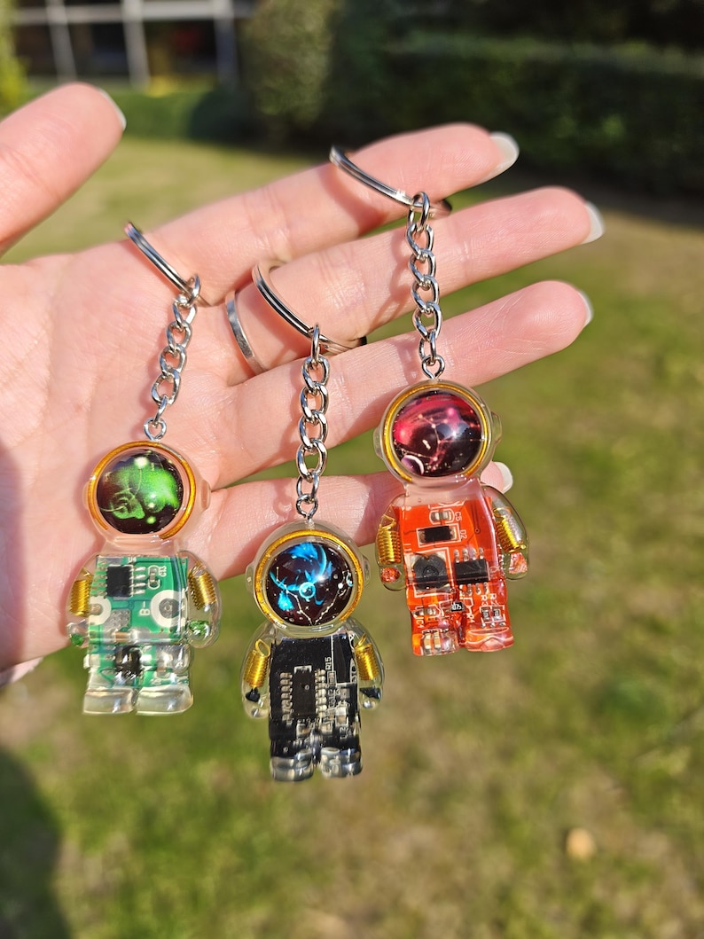 May include: Three astronaut-shaped keychains made of clear resin, each featuring a unique internal circuit board design in green, black, and orange. Each figure has a metallic helmet visor and is attached to a silver-toned metal chain and ring.