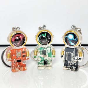 Op de afbeelding: Drie astronautvormige sleutelhangers in oranje, groen en zwart. Elke astronaut heeft een heldere helm met een kleurrijk, galaxy-achtig ontwerp. De sleutelhangers hebben zilverkleurige metalen ringen.