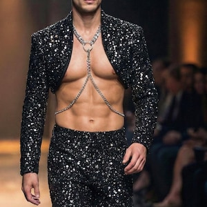 May include: A black sequined bolero jacket and matching pants. The jacket is cropped, and the outfit is accessorized with a silver chain harness. The model is walking on a runway.