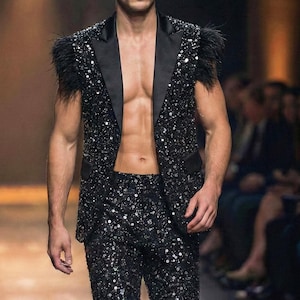 May include: A black sequined suit with a sleeveless jacket featuring feather accents and matching pants. The suit is worn open, revealing the torso. The model is walking on a runway.