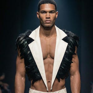 May include: A male model wearing a sleeveless vest with black feather trim and a white lapel. The model is also wearing cream trousers. The background is dark, and the lighting is focused on the model.