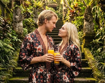 May include: A couple stands on stone steps in a lush, tropical setting, each wearing a black robe with a floral pattern of red and white flowers. They hold glasses of orange juice, with a backdrop of greenery and stone statues.