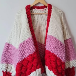 Pode incluir: Um cardigan de malha grossa em creme, rosa e vermelho. O suéter aberto apresenta um design de blocos de cores com um padrão de ponto texturizado nas seções vermelhas. As mangas são uma mistura de rosa e creme. O cardigan é exibido em um cabide de madeira.