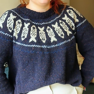 May include: A navy blue sweater with a crew neck and a pattern of white fish around the yoke. The sweater has a speckled texture and a ribbed collar, cuffs, and hem. The sweater is made of a thick, warm material.