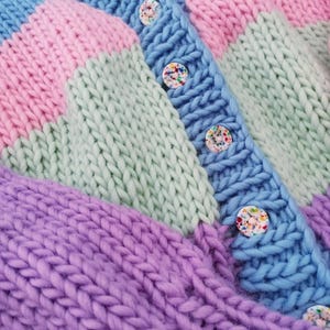 May include: A close-up of a chunky knit cardigan in pastel colors. The sweater features a color-block design with sections of blue, pink, green, and purple. The cardigan has a button-up front with colorful confetti-like buttons.