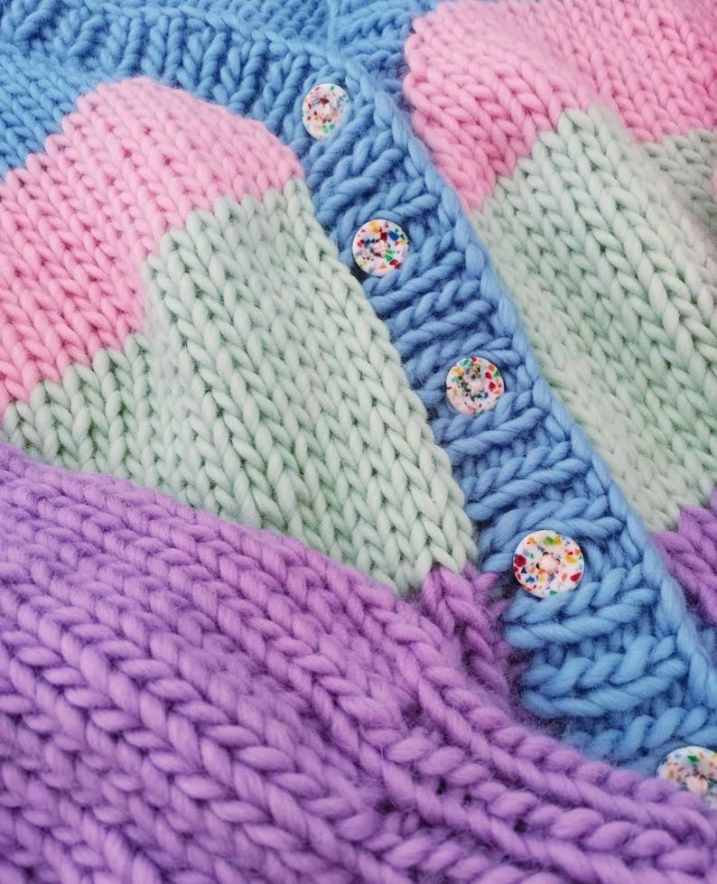 May include: A close-up of a chunky knit cardigan in pastel colors. The sweater features a color-block design with sections of blue, pink, green, and purple. The cardigan has a button-up front with colorful confetti-like buttons.