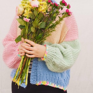 May include: A pastel color block cardigan in pink, peach, green, and blue. The chunky knit sweater has a rainbow trim on the cuffs and hem. The person is holding a bouquet of flowers with pink, yellow, and green stems.