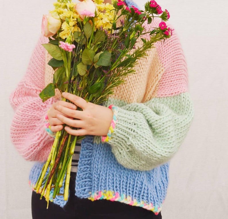 May include: A pastel color block cardigan in pink, peach, green, and blue. The chunky knit sweater has a rainbow trim on the cuffs and hem. The person is holding a bouquet of flowers with pink, yellow, and green stems.