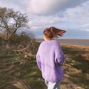 May include: A person wearing a lavender knit sweater and light blue jeans walks outdoors. The sweater is a solid color with a chunky knit texture. The background includes a grassy field, a tree, and a cloudy sky.