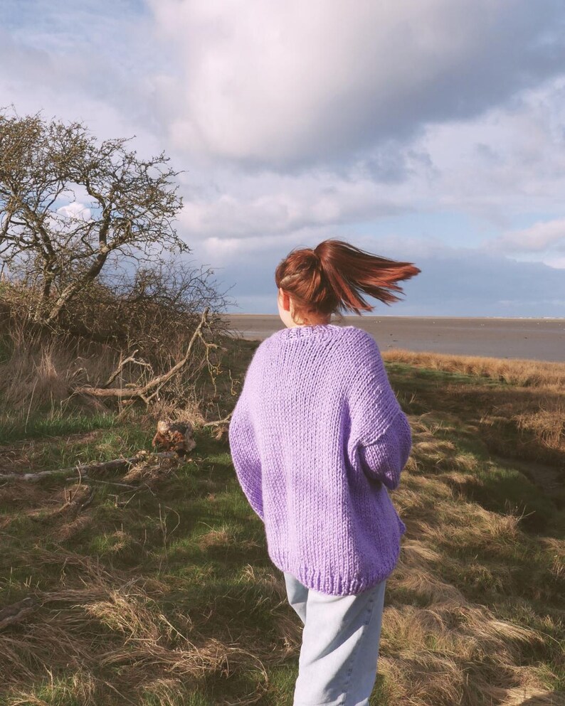 May include: A person wearing a lavender knit sweater and light blue jeans walks outdoors. The sweater is a solid color with a chunky knit texture. The background includes a grassy field, a tree, and a cloudy sky.