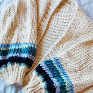 May include: Cream-colored, chunky knit cardigan with blue and teal striped cuffs. The open front sweater features a ribbed texture and a cozy, oversized design. The sleeves have a bold, colorful accent, adding a pop of color to the neutral base.