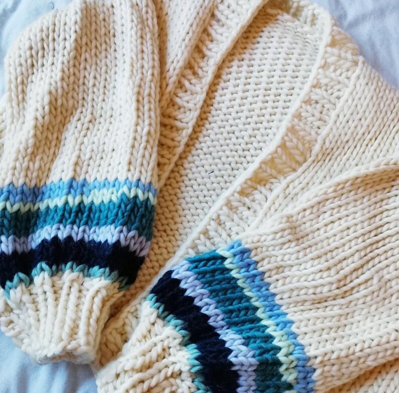 May include: Cream-colored, chunky knit cardigan with blue and teal striped cuffs. The open front sweater features a ribbed texture and a cozy, oversized design. The sleeves have a bold, colorful accent, adding a pop of color to the neutral base.