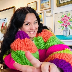 May include: A person wearing a chunky knit sweater with vibrant horizontal stripes of orange, green, pink, and purple. The sweater has a wide, oversized fit. The person is smiling and giving a peace sign.
